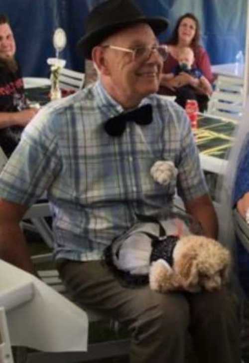 A smiling elderly man in a plaid shirt, bow tie, and hat sits at a table with his small dog dressed in a black-and-white outfit resting on his lap. They appear to be at a celebratory event, surrounded by people and decorations.