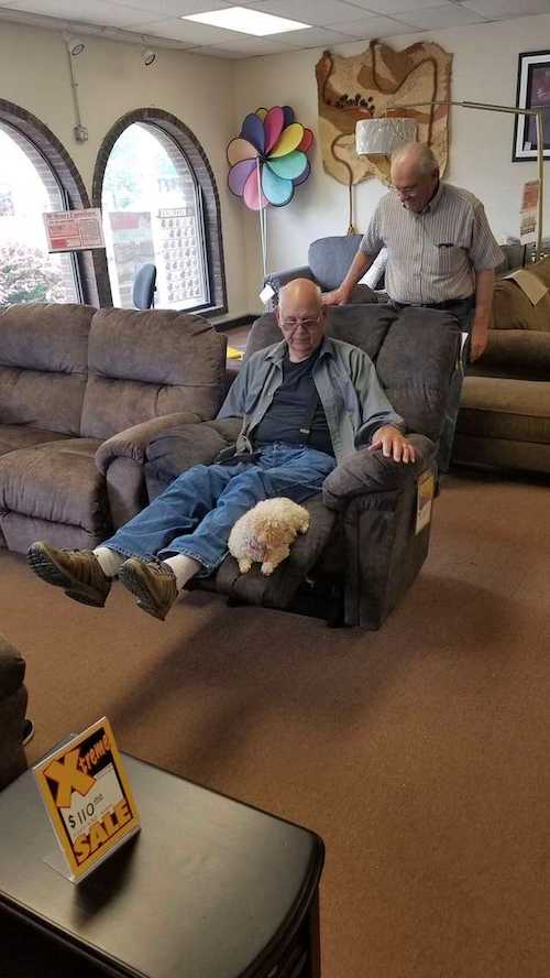 In a cozy furniture store, an elderly man tests out a recliner while his little dog lies contentedly on the extended footrest. Another man stands nearby, watching as they both assess the chair. Sale signs and comfy furniture fill the background.