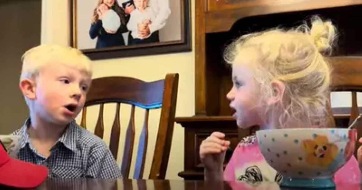 A young blonde boy and a blonde girl are sitting at a dining table, talking animatedly. A colorful bowl with a dog illustration is in front of the girl. Behind them is a family portrait on the wall.