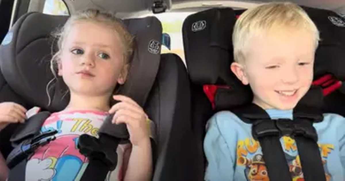 A young boy and girl are sitting side-by-side in car seats. The girl looks serious while adjusting her seatbelt, and the boy smiles brightly, looking pleased.