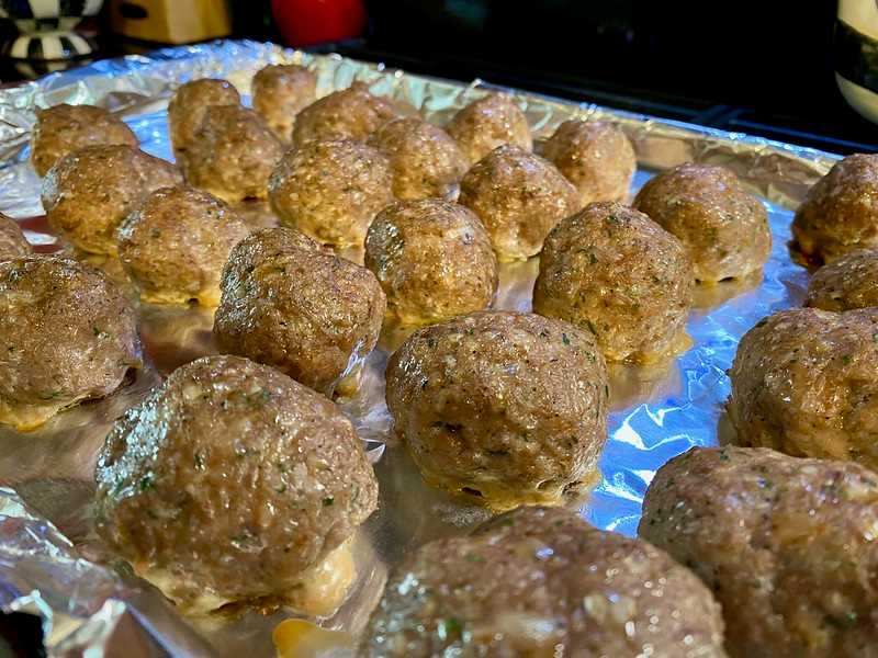 A tray of large, round meatballs arranged in rows on a foil-lined baking sheet, with cheese slightly melting out from underneath some of them.
