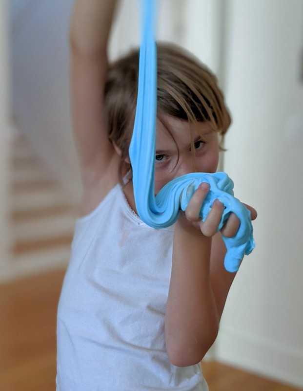 A child in a white tank top stretches a long strand of bright blue slime while holding it close to their face with a playful expression.