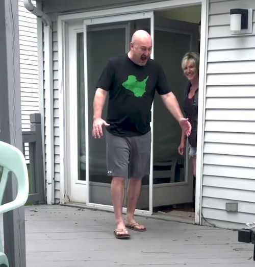 A bald man in a black t-shirt and shorts reacts with wide eyes and open arms upon seeing something on the porch, while a woman peeks out from the doorway behind him.