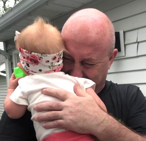 A man holds a baby close to his face with his eyes shut and tears visible, while the baby wears a floral headband and white shirt.