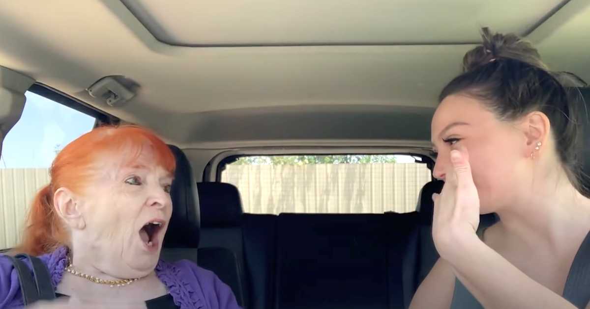 An elderly woman with bright red hair sits in the passenger seat of a car, reacting with joyful surprise while looking at a young woman in the driver's seat, who is smiling and covering her mouth in excitement.