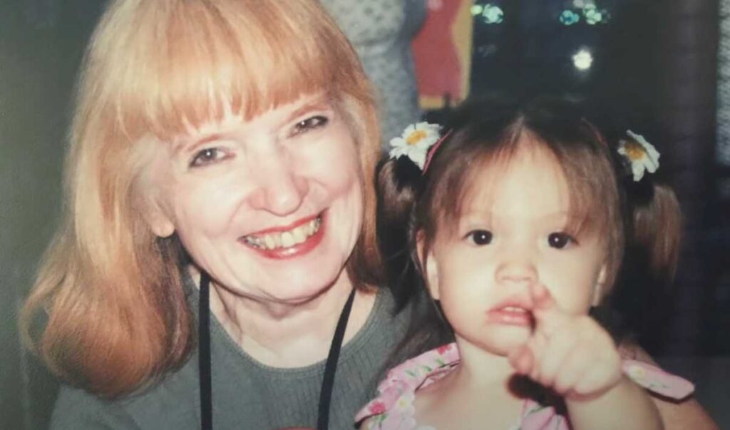 A smiling elderly woman with light red hair poses with a young girl who has pigtails and small white flowers in her hair; the girl is pointing toward the camera with a curious expression.