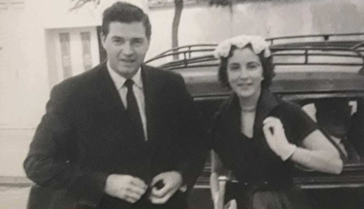 A cropped, closer version of the previous photo, showing the couple from the waist up. The man has short dark hair and is dressed in a dark suit. The woman has short curly hair, is wearing gloves, and has a soft smile.