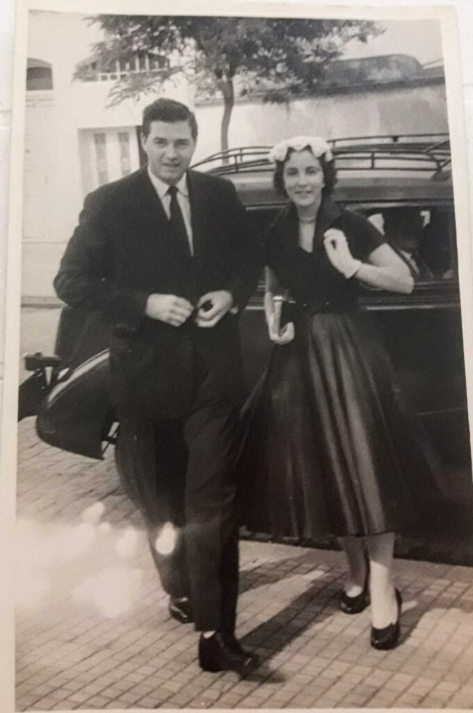 A black-and-white photo of a young couple standing outside in front of a classic car. The man is in a suit and tie, and the woman is wearing a full-skirted dress with a headband or decorative hat. They both appear to be smiling.