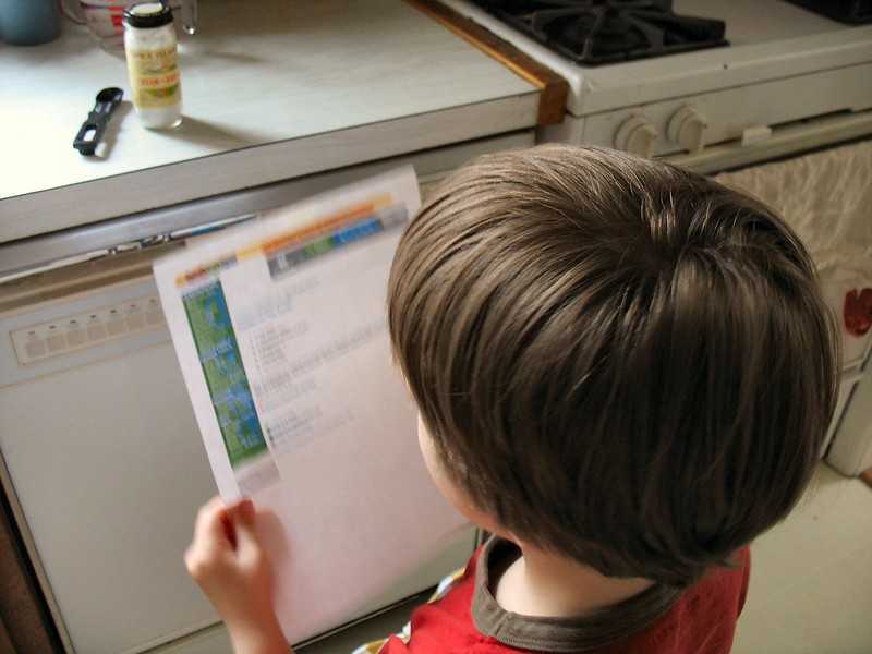 A young boy stands in a kitchen holding a printed recipe sheet, appearing to read instructions.