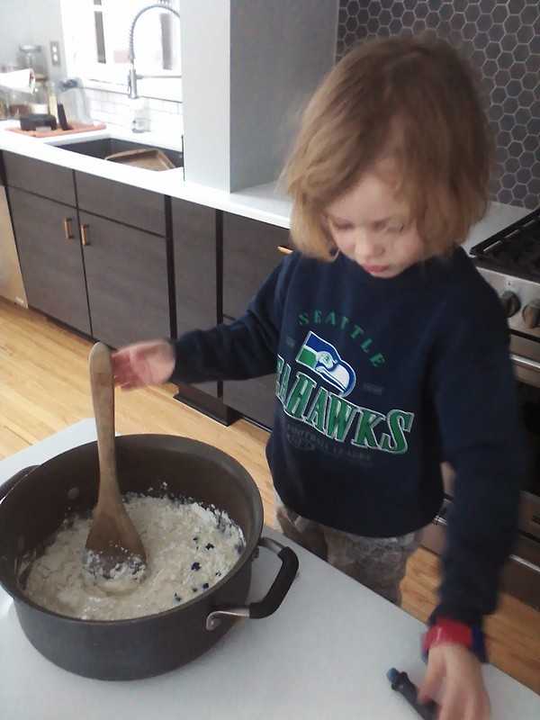 A child in a Seahawks sweatshirt stirs a large black pot of homemade playdough on a kitchen counter.