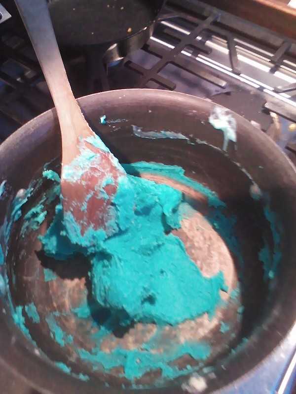 A wooden spoon mixes a thick, teal-colored dough inside a black cooking pot on a stove.