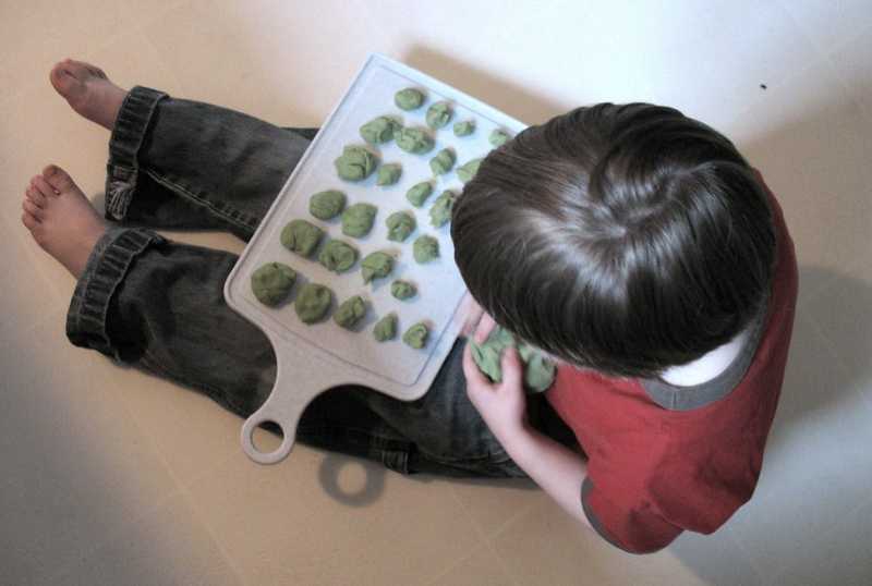 A young child sits on the floor with a cutting board full of small green playdough balls, shaping one by hand.