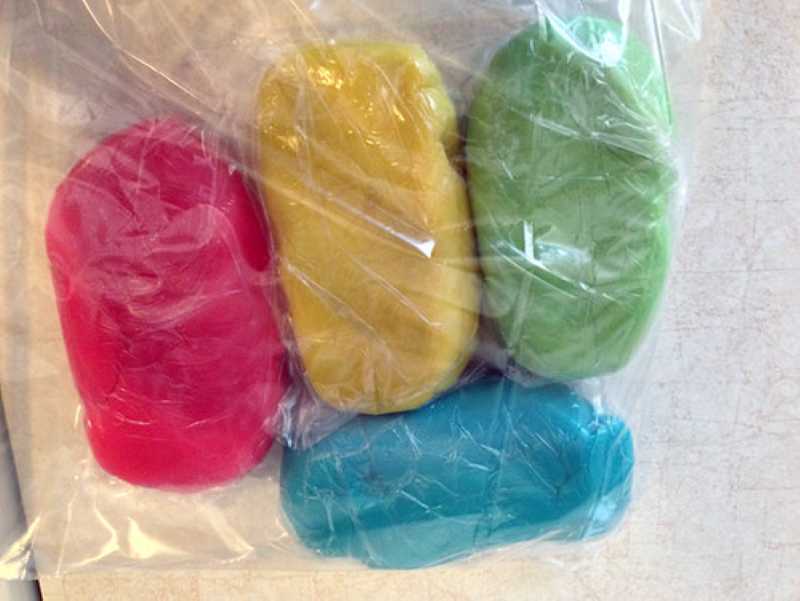 Four brightly colored lumps of homemade playdough—pink, yellow, green, and blue—are wrapped in plastic and placed on a counter.