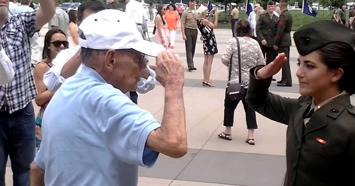An elderly man in a light blue polo shirt and white cap salutes a young woman in a Marine uniform. They face each other outdoors on a paved area, surrounded by a crowd of people, some also saluting. The moment appears emotional and ceremonial, likely a military event or graduation.