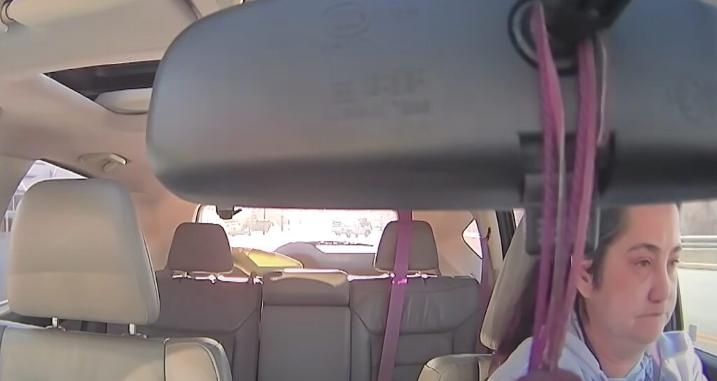 A woman with a serious expression is seen driving a vehicle, captured from a dashcam perspective inside the car, with the rearview mirror and hanging decorations partially obscuring the view. The backseat and rear window are visible in the bright daylight.
