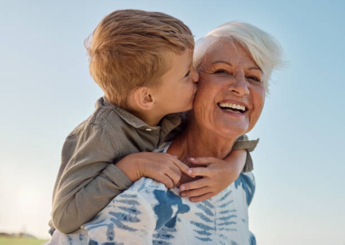 A young boy kisses his smiling grandmother on the cheek while hugging her from behind. The grandmother, with short white hair, is wearing a blue and white patterned top, and they are both outdoors under a clear blue sky.