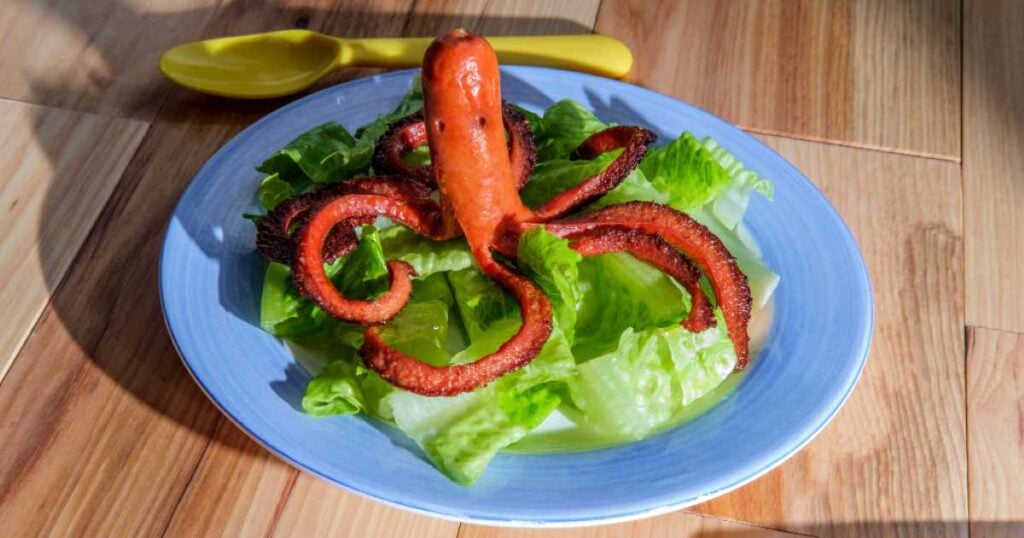 A creatively cut and cooked hot dog shaped like an octopus with curled tentacles, sitting on a bed of romaine lettuce on a blue plate.