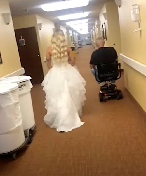 A rear view of a bride walking through a nursing home hallway, her long blonde hair flowing over her white gown, approaching a seated man in a wheelchair.