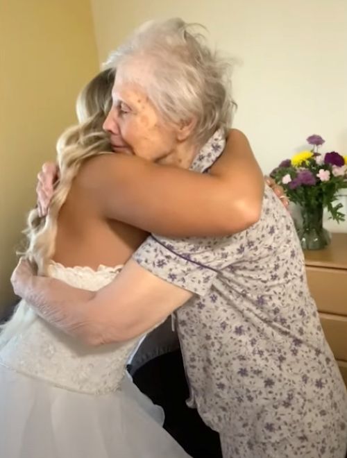 An elderly woman in a purple floral top embraces her granddaughter, dressed as a bride, beside a small table with a vase of flowers.