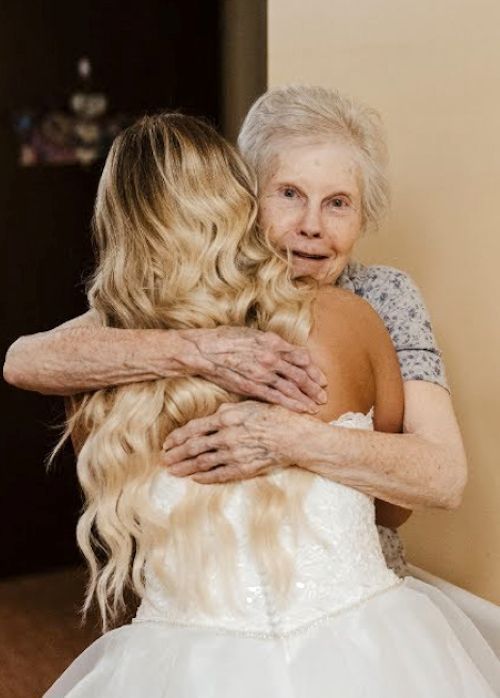 An elderly woman looks joyfully at the camera while hugging her granddaughter, who is in a white wedding dress, showing a moment of deep connection.