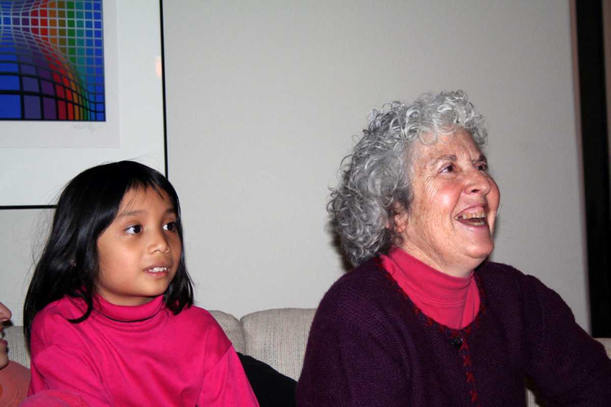 A smiling grandmother with curly gray hair sits beside her curious young granddaughter, both wearing pink and purple tops, sharing a joyful moment indoors—capturing the heartwarming bond that defines being a grandmother.