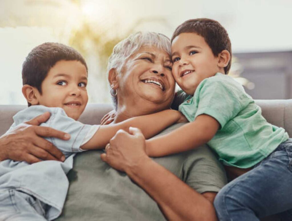 Smiling grandmother sitting on a couch while being hugged tightly by two young grandsons, both wearing casual clothes and looking happy and playful.