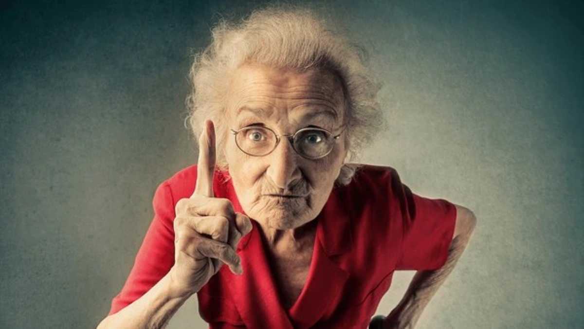 Elderly woman with white hair, glasses, and a serious expression wearing a red shirt, pointing her index finger upward in a scolding or warning gesture, standing against a neutral background.