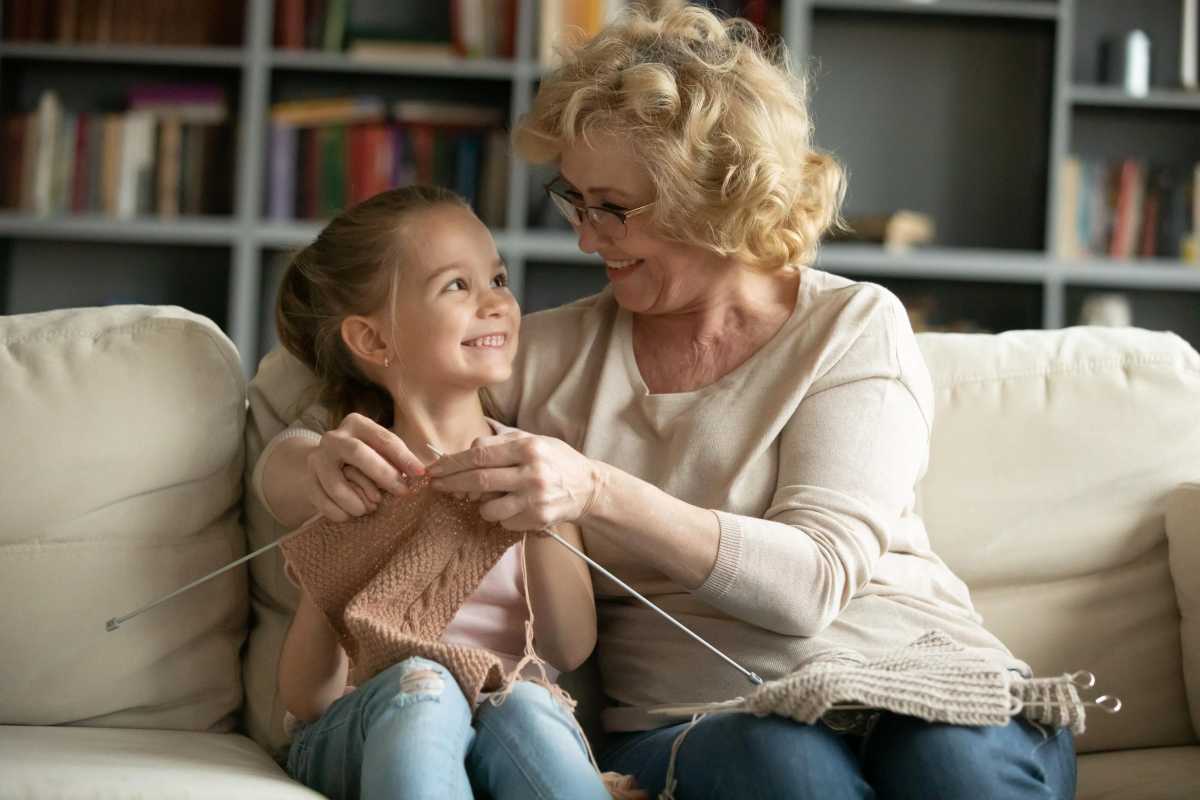 A smiling grandmother teaches her granddaughter how to knit as they sit together on a cozy couch, enjoying a warm and loving moment.