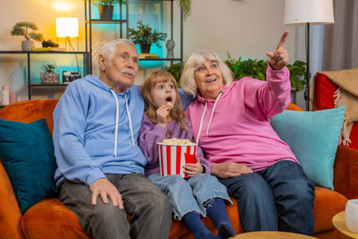 An elderly man, a young girl, and an elderly woman sit together on an orange couch, watching something with interest. The man wears a light blue hoodie, the girl wears a purple sweater and holds a large bucket of popcorn, and the woman wears a pink hoodie while pointing excitedly toward the screen. All three appear engaged and happy in a cozy, well-lit living room with plants and colorful decor in the background.