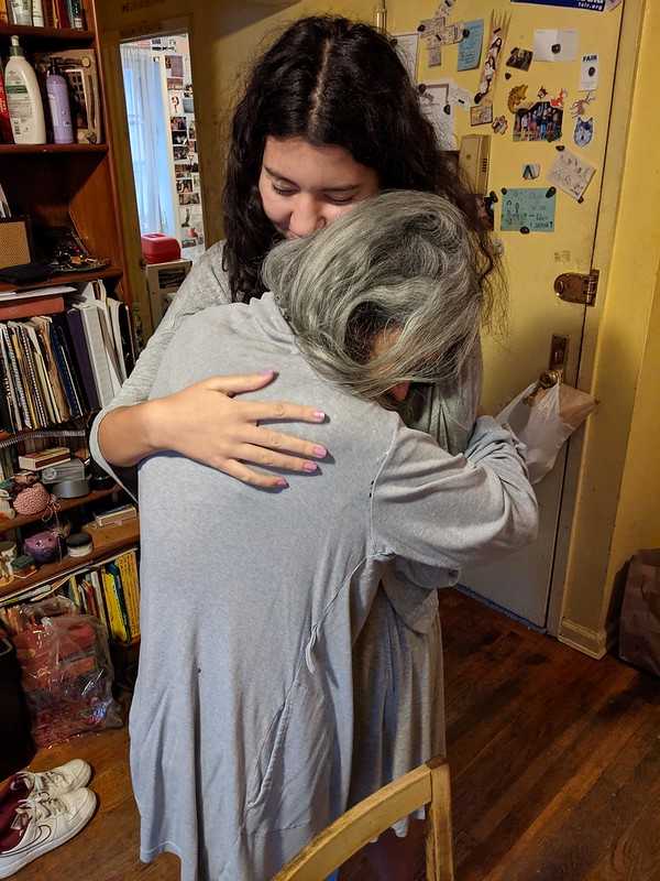 A grandmother with gray hair hugging a young woman tightly in a cozy, cluttered home, sharing an emotional and loving moment.