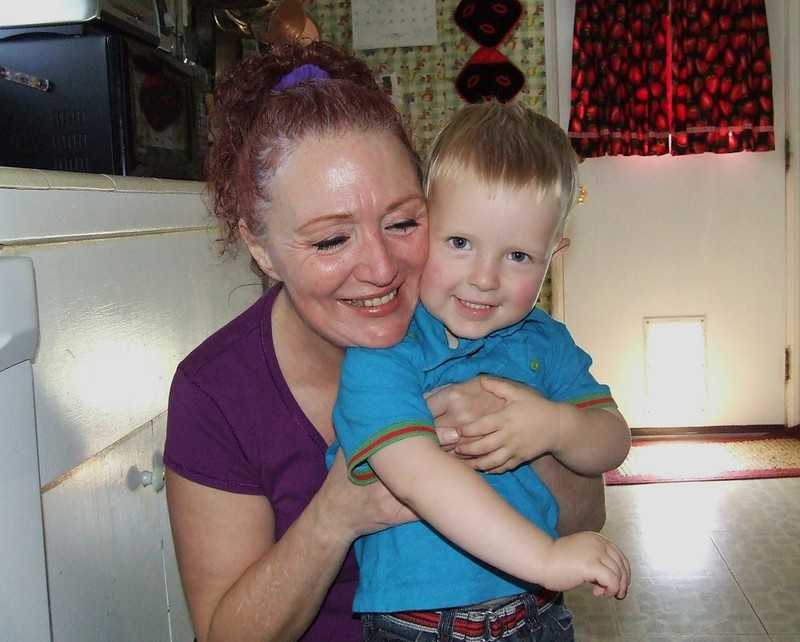 These 67 Open-Ended Questions Are the Golden Keys to Your Grandchild’s Heart 2 A joyful grandmother with red hair in a ponytail hugs her smiling young grandson in a cozy kitchen with patterned wallpaper and red curtains.