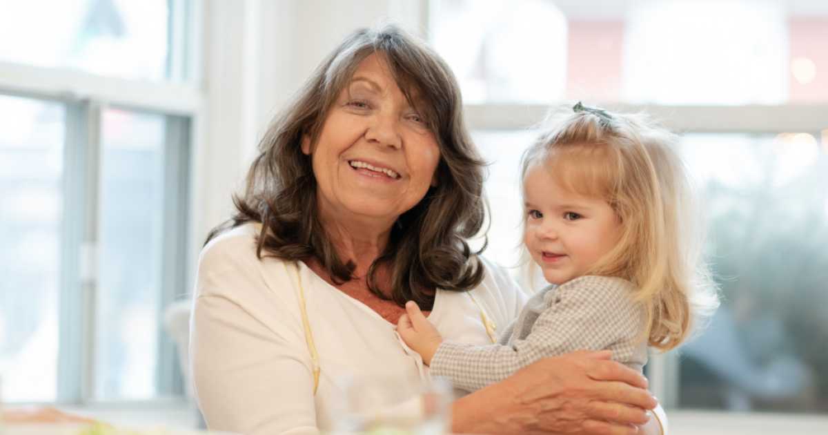 A smiling grandmother with long brown hair holds her young granddaughter in her arms inside a bright room with large windows in the background.