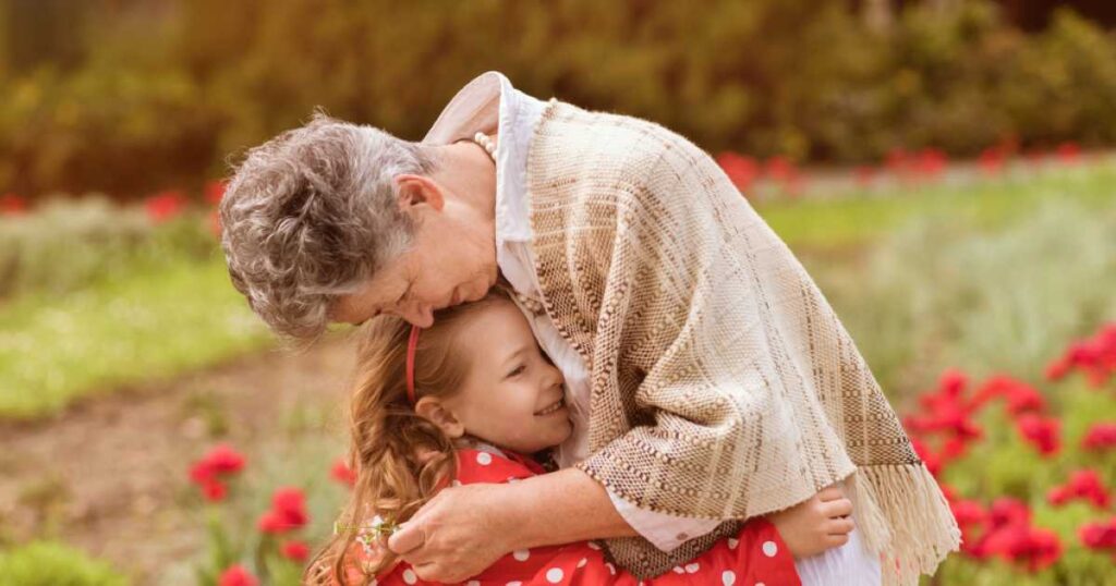 An elderly woman with short gray hair lovingly hugs her granddaughter in a red polka dot dress while standing in a flower garden filled with red blossoms.