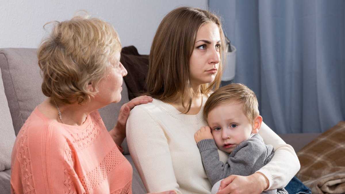 An older woman sits beside a younger woman holding a small child on her lap. The older woman appears to be talking gently, while the younger woman looks upset and holds the child protectively. The child looks serious and clings closely to the younger woman.