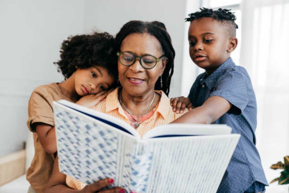 An older woman wearing glasses reads a book to two young children, one resting their head on her shoulder and the other leaning in closely, all appearing engaged and happy together.