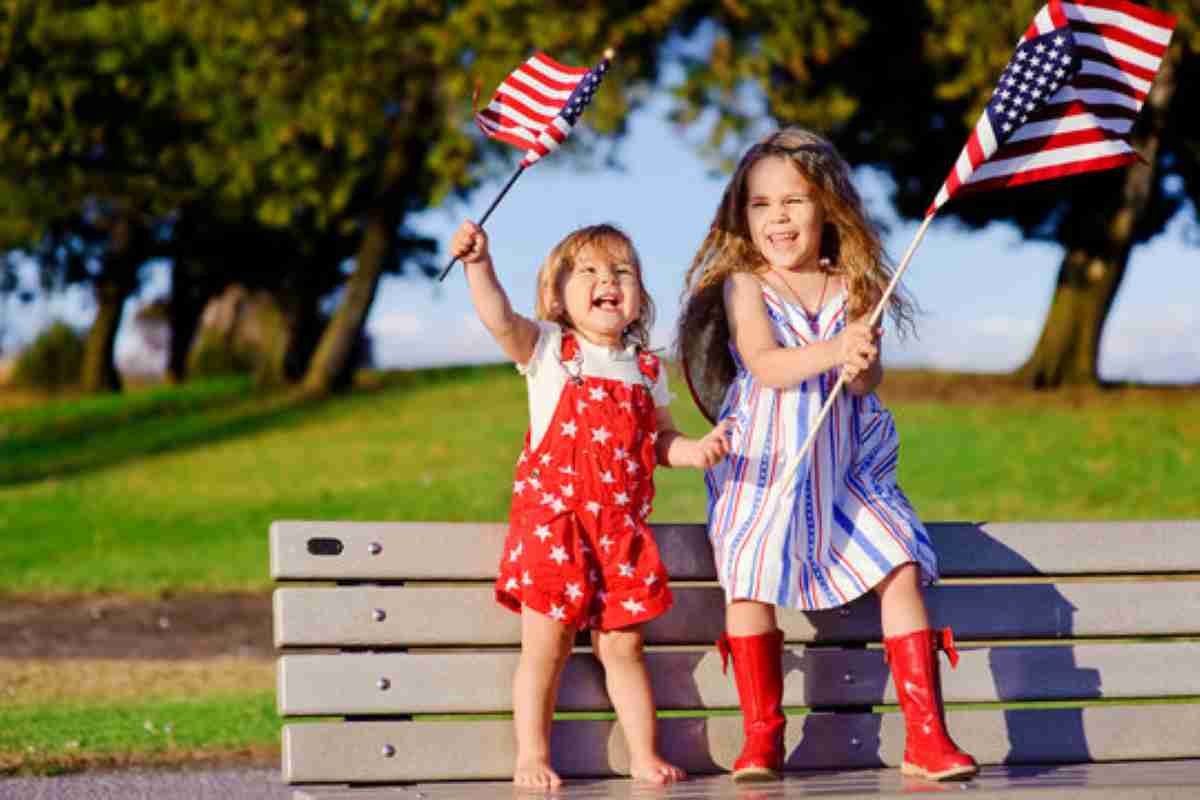 Two young girls celebrate the 4th of July while sitting on a park bench. The younger girl stands barefoot in red overalls with white stars and waves a small American flag. The older girl wears a red, white, and blue striped dress with red boots, holding a larger American flag. Both are smiling and appear joyful, with green grass and trees in the background.