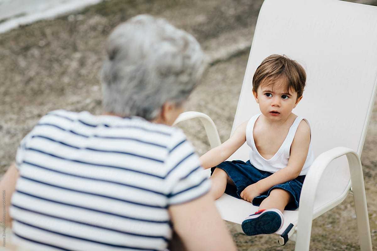 A young boy wearing a white tank top and blue shorts sits on a white outdoor chair, looking attentively at an elderly woman with short gray hair wearing a striped shirt who is facing him.