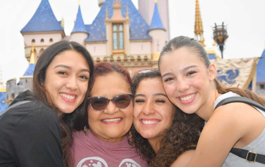 A heartwarming moment at Disneyland featuring an older woman in sunglasses smiling joyfully while hugging three young women, all beaming with excitement, with the iconic Disney castle in the background.