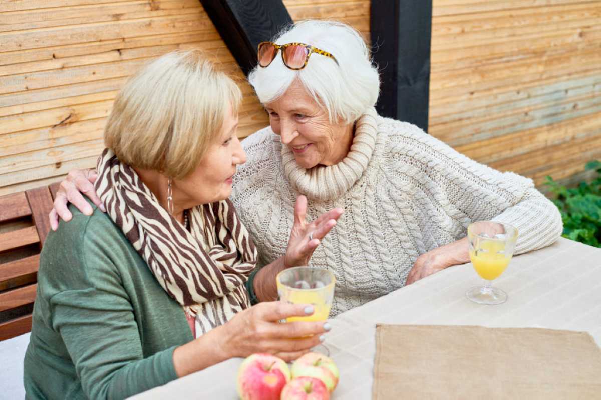Two elderly women sit closely together outdoors, smiling and talking warmly over glasses of juice, with apples on the table—capturing a heartfelt moment of friendship and shared advice between grandmas.