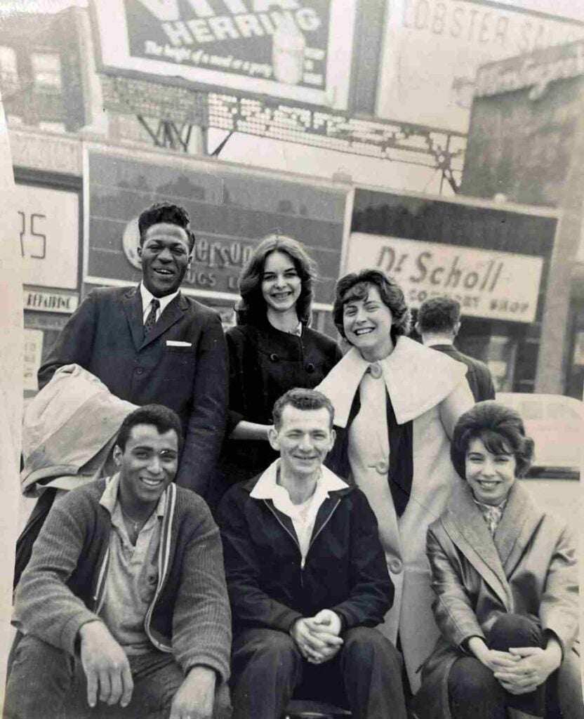 A black-and-white photo of six friends smiling and posing in a busy city area with storefront signs behind them.
