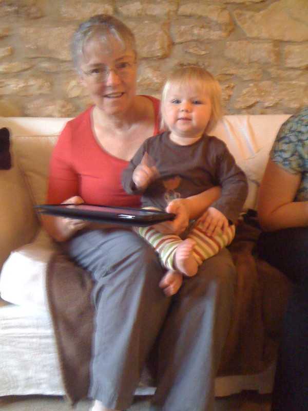 An older woman with short gray hair sits on a couch with a smiling toddler on her lap. The woman is holding a closed laptop while the toddler, dressed in a brown shirt and striped pants, looks at the camera with one hand raised. A stone wall serves as the cozy background.