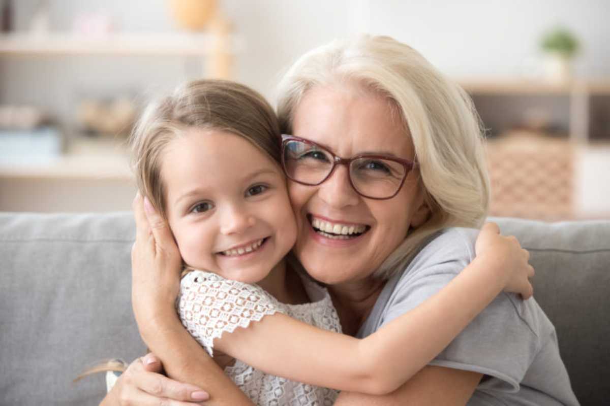 A joyful grandmother with long silver-blonde hair and glasses hugs her young granddaughter on a couch. Both are smiling brightly, their faces close together, radiating warmth and happiness. The little girl wears a white lace top, and the background is softly blurred with homey decor.