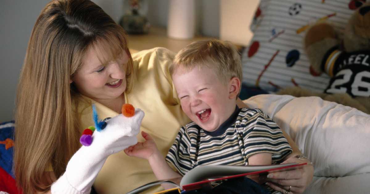 A smiling woman holding a sock puppet decorated with colorful pom-poms, making a young boy laugh while they read a book together.