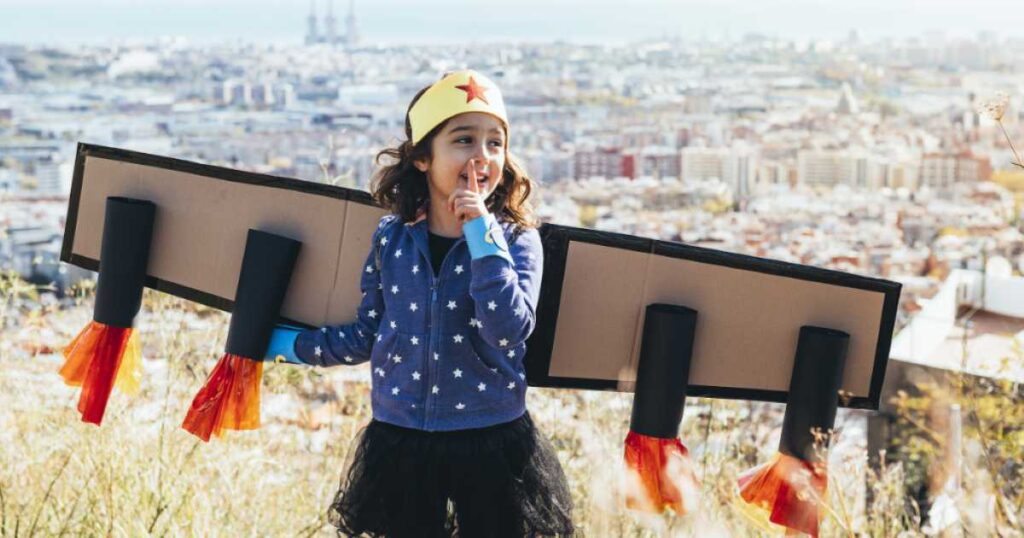 A young girl wearing a cardboard rocket costume and a yellow crown, smiling with her finger to her lips while standing on a hill overlooking a city.