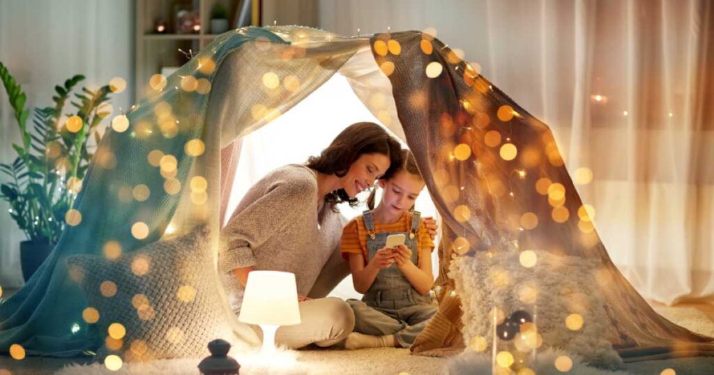 A woman and a young girl sitting inside a cozy blanket fort with string lights, smiling as they look at a smartphone together.