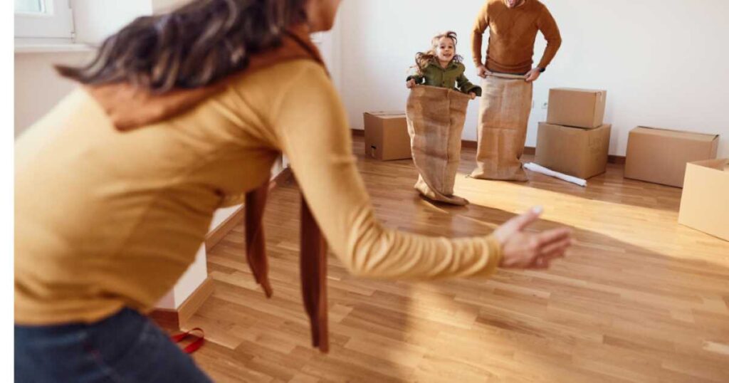 A family playing a sack race indoors, with a child hopping toward an adult reaching out with open arms.