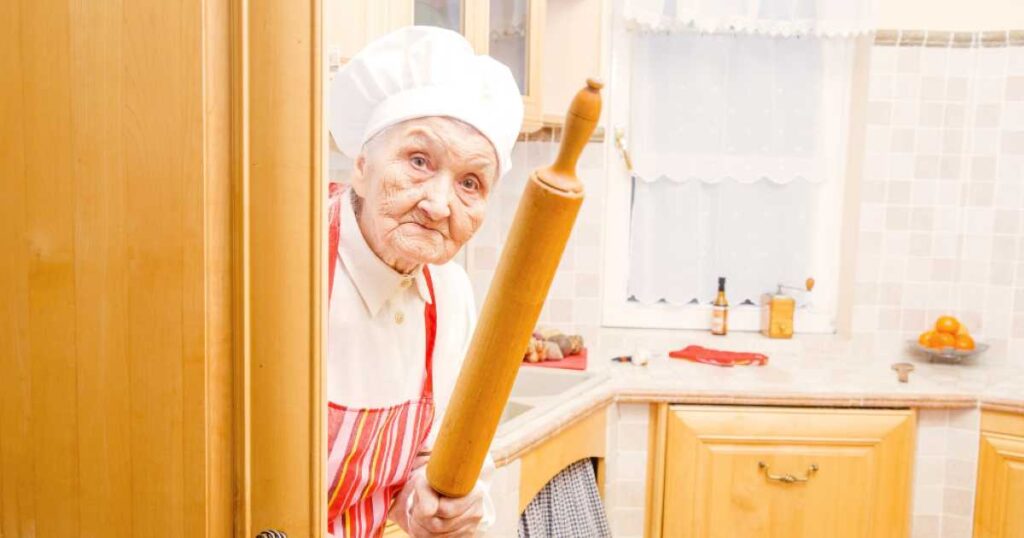 An elderly woman in a chef hat and apron peeking from behind a kitchen door, holding a rolling pin with a playful expression.