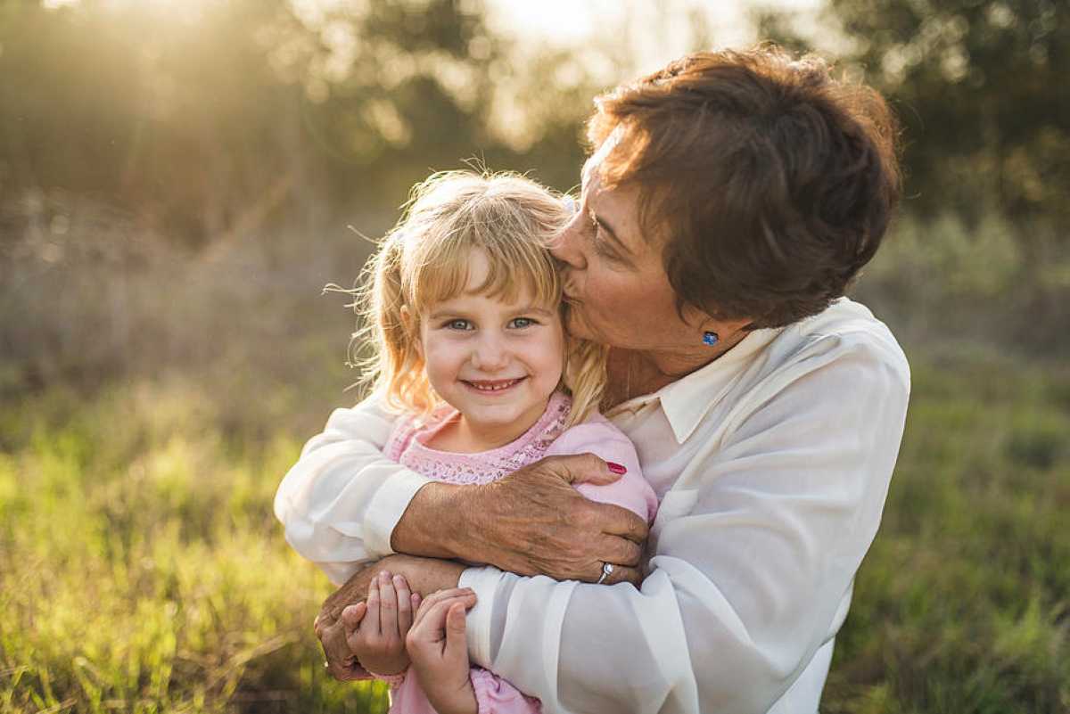 An older woman lovingly hugs and kisses a smiling young girl on the cheek while standing outdoors in a sunny field. The grandmother wears a white blouse, and the little girl, dressed in a pink top, looks directly at the camera with a happy expression. Warm sunlight filters through the background, creating a soft and heartwarming scene.