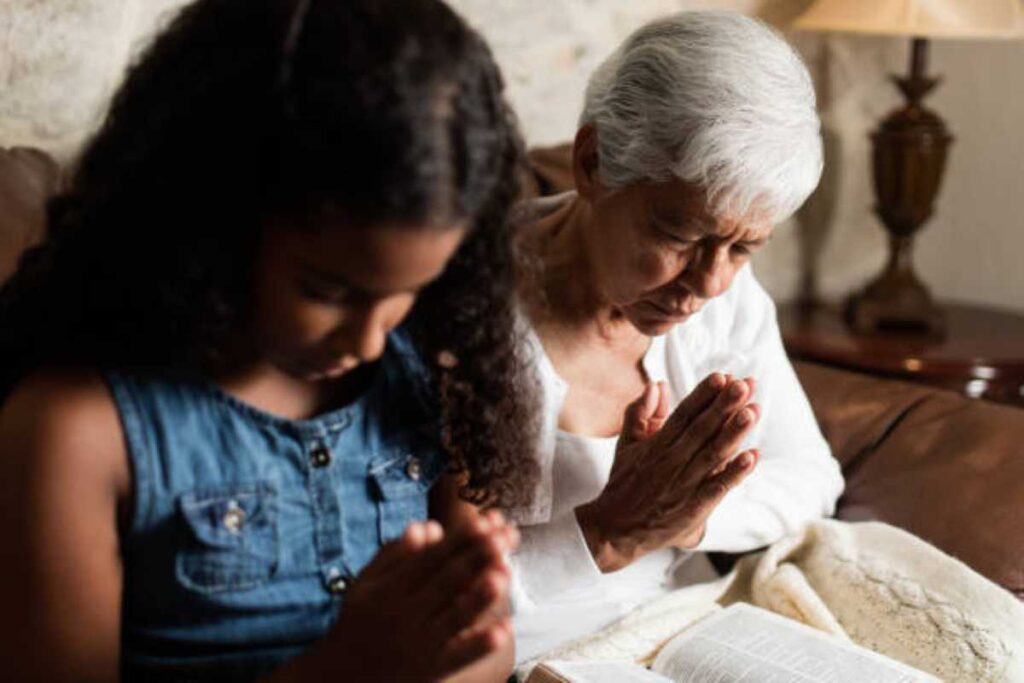 An elderly woman with short white hair and a young girl with long curly hair sit side by side on a couch with their heads bowed and hands clasped in prayer. A Bible rests open on their laps, and a cozy blanket is draped over them. The setting is warm and peaceful, lit by a lamp in the background.