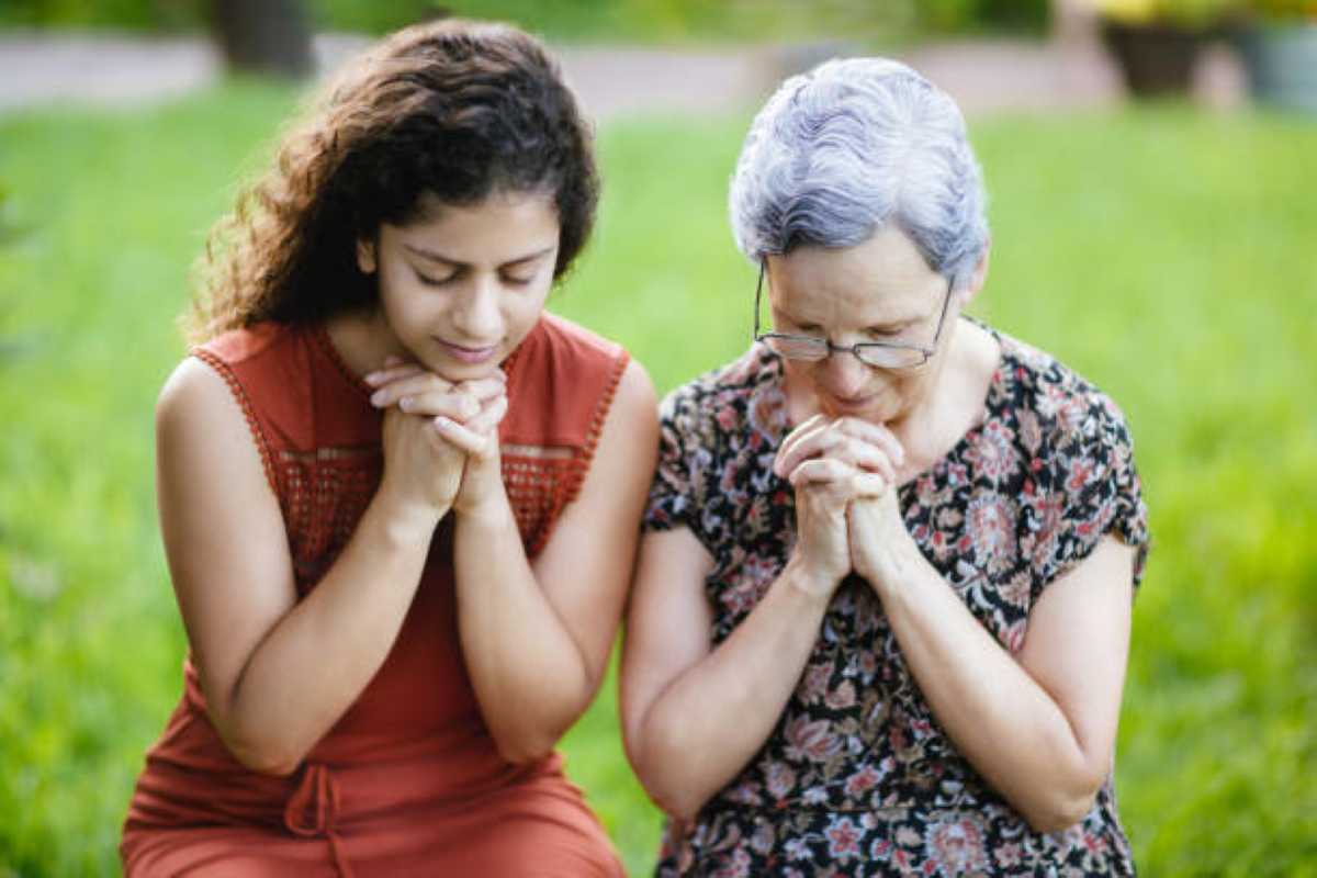 A grandmother with short gray hair and glasses kneels on the grass beside her granddaughter, both with heads bowed and hands clasped in prayer. The girl wears a sleeveless rust-colored dress, and the older woman wears a floral-print dress. They are outdoors in a serene, green park setting.